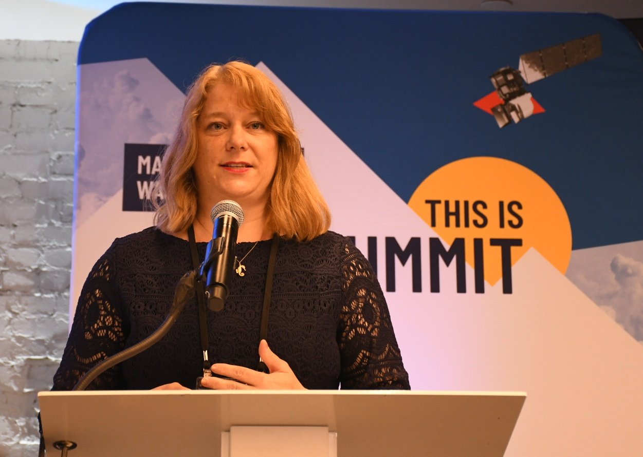 Amy Larson stands behind a podium in front of the SatSummit banner.