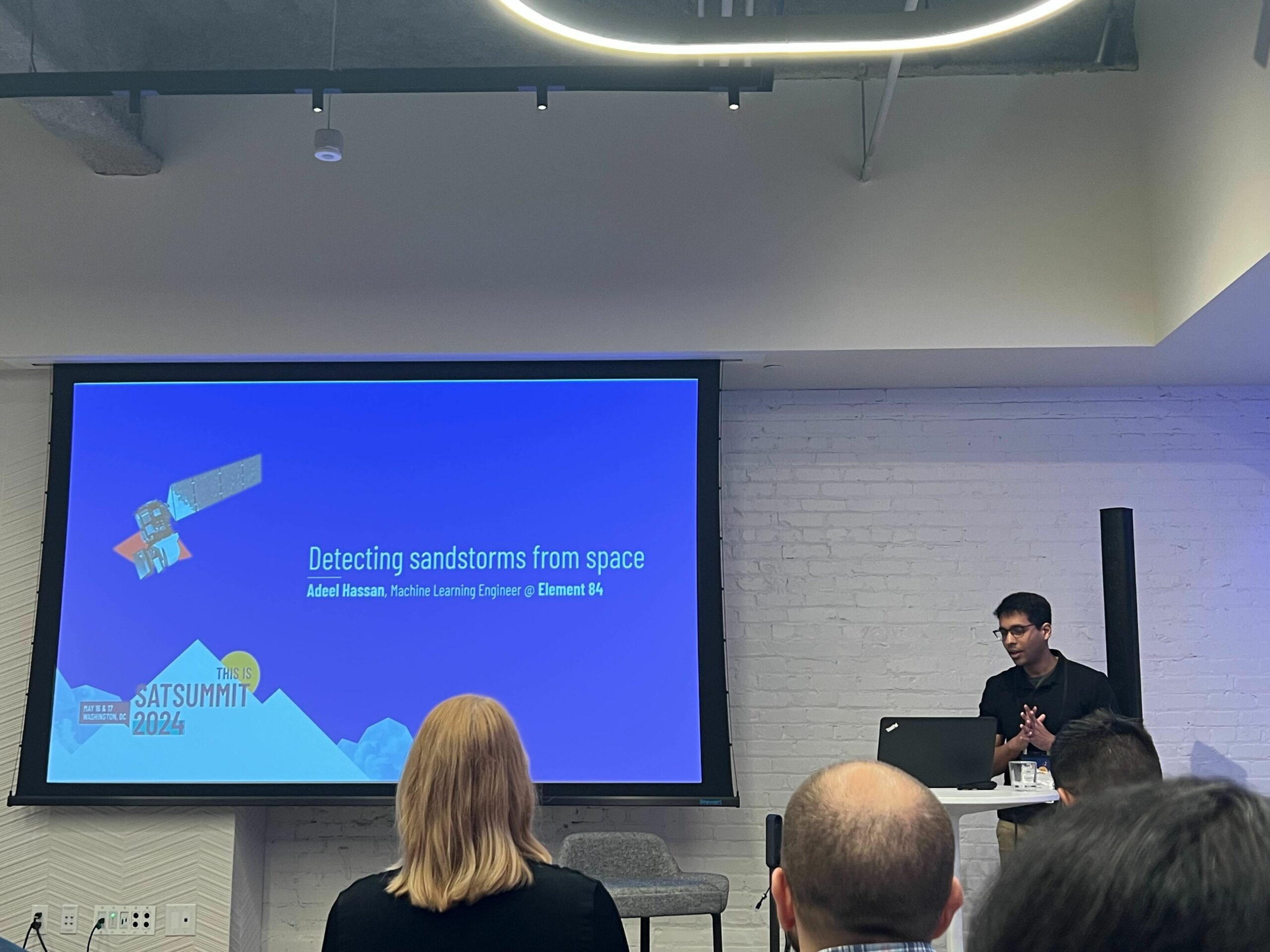 Adeel Hassan stands behind a podium next to a projector screen that reads, "Detecting sandstorms from space" and his name, position title, and Element 84.