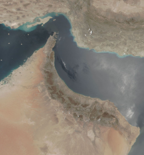 An aerial image depicting desert terrain and water. There are flecks of either dust or reflected sunlight over the water.